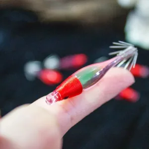Red green mini transparent shrimp squid jig held on finger showing compact size with red cloth head and crown hooks